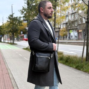 以下が含まれることがあります： シルバーのジッパー開閉と調節可能なストラップが付いた、黒い革のクロスボディバッグ。長方形のバッグはミニマリストなデザインです。バッグはグレーのコートの上から着用されています。