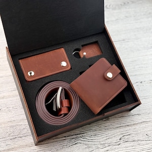 Könnte beinhalten: Ein braunes Leder-Geschenkset in einer Holzbox. Das Set enthält ein Portemonnaie, einen Kartenhalter, einen Gürtel und einen Schlüsselanhänger. Der Gürtel hat eine silberne Schnalle. Die Artikel sind in einem schwarzen Schaumstoffeinsatz angeordnet.