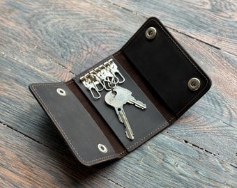 Könnte beinhalten: Offener, dunkelbrauner Leder-Schlüsselhalter mit silbernen Schlüsselringen und zwei Schlüsseln. Der Schlüsselhalter ist geöffnet und zeigt das Innere und die Schlüssel. Das Leder hat einen Used-Look und der Schlüsselhalter wird mit Druckknöpfen verschlossen.