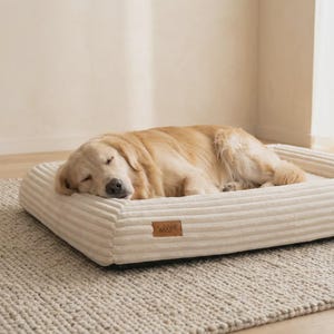 Puede incluir: Un golden retriever durmiendo sobre una cama para perros de pana color crema. La cama tiene forma rectangular con laterales elevados y una pequeña etiqueta de cuero marrón con la palabra "Woope". La cama está sobre una alfombra beige texturizada.