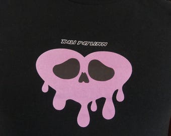 Puede incluir: Camiseta negra con un diseño de calavera rosa con detalles de goteo. El texto "KALI PAYLINN" está impreso encima de la calavera. La camiseta es de cuello redondo.