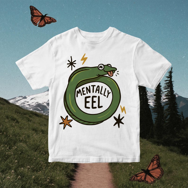 May include: White t-shirt featuring a cartoon eel design. The green eel is coiled with the words "MENTALLY EEL" inside. The design includes lightning bolts and stars. Two orange butterflies are in the background.