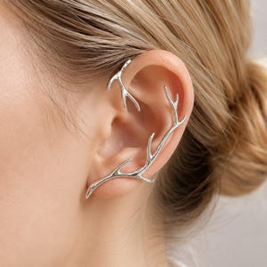 May include: A silver-toned ear cuff designed to resemble branching antlers. The piece features two distinct sections that follow the natural curve of the ear, with one smaller branch resting at the top and a larger, elongated branch along the side.
