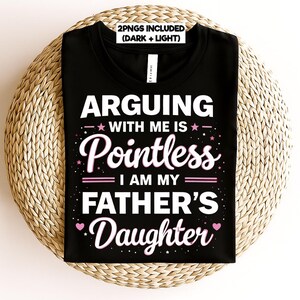 May include: Black t-shirt with the text "ARGUING WITH ME IS Pointless I AM MY FATHER'S Daughter" in white and pink lettering. The shirt is displayed on a woven, circular surface.