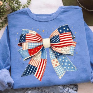 Peut inclure: Un sweat-shirt bleu clair avec un grand motif de nœud. Le nœud est fait de tissu patchwork avec des motifs rouges, blancs et bleus, y compris des étoiles et des rayures. Des étoiles dorées sont dispersées autour du nœud.