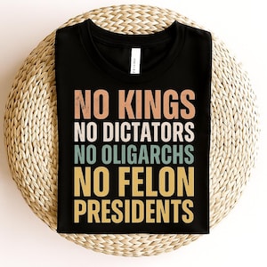 Puede incluir: Camiseta negra con el texto "NO KINGS NO DICTATORS NO OLIGARCHS NO FELON PRESIDENTS" en letras naranjas, blancas, verde azulado y amarillas. La camiseta se muestra sobre una superficie redonda y tejida.