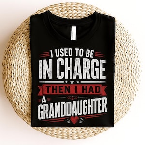 May include: Black t-shirt with white and red text that reads "I USED TO BE IN CHARGE THEN I HAD A GRANDDAUGHTER" with a heart graphic. The shirt is displayed on a woven, circular surface.