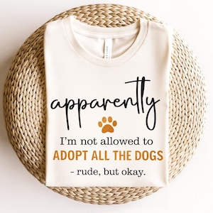 Può includere: T-shirt color crema con la parola "apparently" in carattere scritto a mano, un'impronta di zampa e la frase "I'm not allowed to ADOPT ALL THE DOGS - rude, but okay." La maglietta è esposta su un tappetino circolare intrecciato.