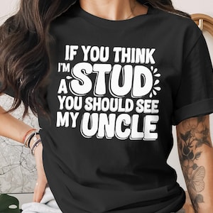 Peut inclure: T-shirt noir avec texte blanc : « IF YOU THINK I'M A STUD YOU SHOULD SEE MY UNCLE ». Le texte est en gras et en police fantaisie. Le t-shirt a des manches courtes et un col rond.