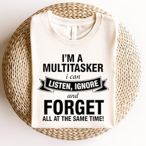 Puede incluir: Camiseta color crema con el texto negro: "I'M A MULTITASKER i can LISTEN, IGNORE and FORGET ALL AT THE SAME TIME!". La camiseta se muestra sobre una superficie circular tejida.