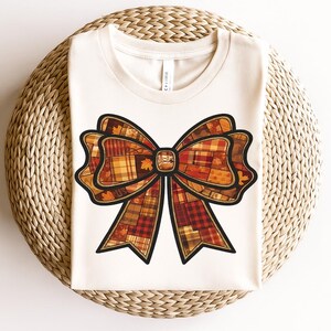 Peut inclure: T-shirt crème avec un grand motif de nœud. Le nœud est un patchwork de couleurs automnales, dont le rouge, l'orange et le jaune, avec un contour noir. Le motif comprend des accents de feuilles, parfait pour l'automne.