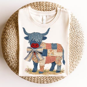 May include: Cream-colored t-shirt featuring a patchwork Highland cow design. The cow has blue denim horns and a denim head, a red nose, and a plaid bow tie. The body is made of various patterned fabrics.