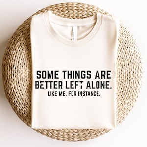 Puede incluir: Camiseta color crema con el texto "SOME THINGS ARE BETTER LEFT ALONE. LIKE ME, FOR INSTANCE." en letras negras en negrita. La camiseta está doblada y colocada sobre una superficie circular tejida.
