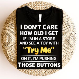 Peut inclure: T-shirt noir avec texte blanc : "I DON'T CARE HOW OLD I GET IF I'M IN A STORE AND SEE A TOY WITH 'Try Me' ON IT, I'M PUSHING THOSE BUTTONS". Le t-shirt est présenté sur une surface tissée.