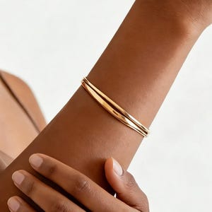 Puede incluir: Una pulsera dorada con un acabado liso y pulido. La pulsera tiene un diseño simple y elegante, y parece estar hecha de metal. La pulsera se lleva en el brazo.