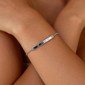 May include: A silver bracelet with a rectangular bar pendant. The bar is engraved with the text "XI • LYNN VI". The bracelet is on a delicate chain.