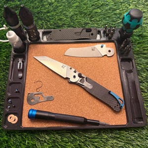May include: A black tray with a corkboard center holds various tools and knives. The tray includes a screwdriver, multiple knife blades, and small parts. The knives have silver blades and black and blue handles. Several bottles are also in the tray.