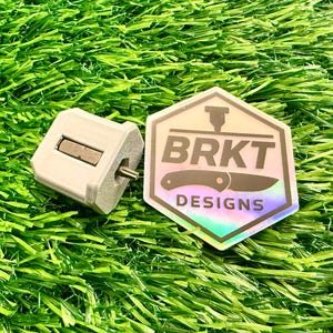 May include: A silver-colored knife sharpener and a holographic sticker with the text "BRKT DESIGNS" and a knife graphic. The items are on a bed of green artificial grass.