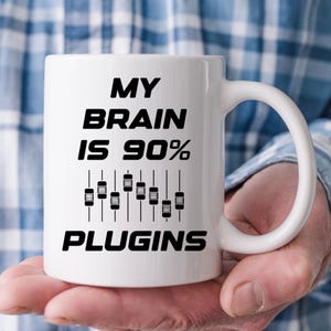 May include: White ceramic mug with the text "MY BRAIN IS 90% PLUGINS" in black, and a graphic of equalizer sliders. The mug has a curved handle. The mug is held by a person.