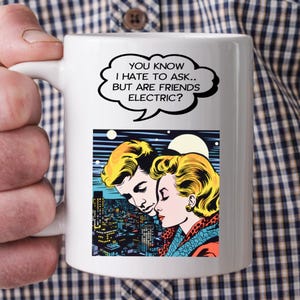 Puede incluir: Taza de cerámica blanca con una ilustración de estilo cómic de una pareja y un paisaje urbano. La taza tiene el texto "YOU KNOW I HATE TO ASK.. BUT ARE FRIENDS ELECTRIC?" en un bocadillo.