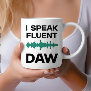 May include: White ceramic mug with the text "I SPEAK FLUENT DAW" in black, with a teal sound wave graphic. The mug has a white handle and is held by a person.