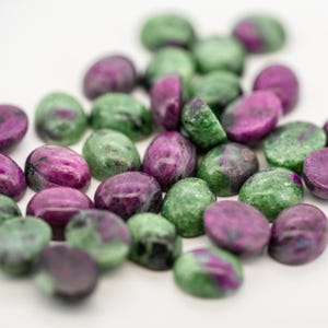 May include: A close-up of a collection of oval gemstones in green and purple hues. The stones are scattered on a white surface, with some cut to reveal their internal patterns. The lighting accentuates the natural colours and textures of the gemstones, creating a visually appealing display.
