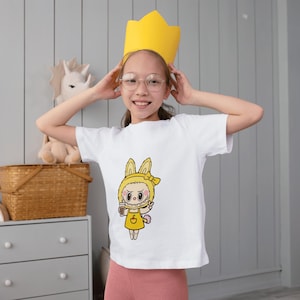 Puede incluir: Una camiseta blanca con un personaje de dibujos animados de conejito amarillo sosteniendo una bebida. El personaje tiene una expresi&oacute;n seria. Una persona joven lleva la camiseta y una corona de papel amarilla.