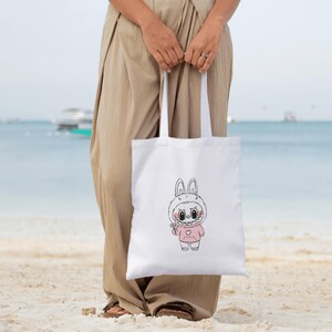 Puede incluir: Bolso tote blanco con un personaje de conejo de dibujos animados con una sudadera con capucha rosa. El conejo tiene una sonrisa con dientes y sostiene una peque&ntilde;a flor. El bolso tiene asas blancas largas y lo sostiene una persona en una playa.