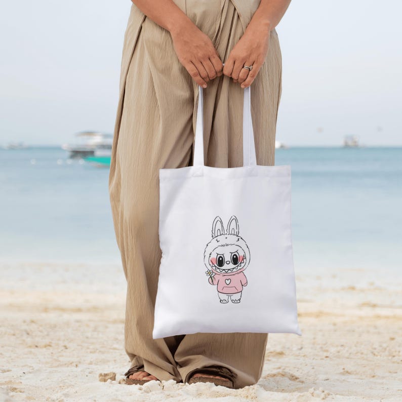 Puede incluir: Bolso tote blanco con un personaje de conejo de dibujos animados con una sudadera con capucha rosa. El conejo tiene una sonrisa con dientes y sostiene una peque&ntilde;a flor. El bolso tiene asas blancas largas y lo sostiene una persona en una playa.
