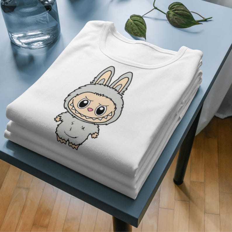 Puede incluir: Una pila de camisetas blancas con un monstruo de dibujos animados con disfraz de conejo. El monstruo es gris con ojos grandes y una sonrisa dentuda. Las camisetas est&aacute;n cuidadosamente dobladas sobre una superficie azul.