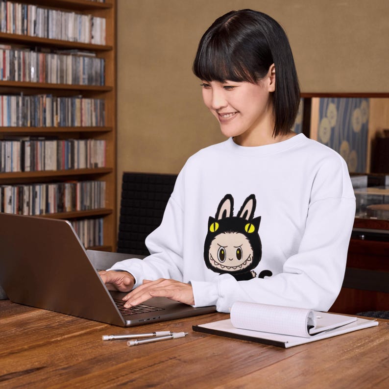 Puede incluir: Una sudadera blanca con un personaje de gato de dibujos animados con un disfraz negro con ojos amarillos y una sonrisa dentuda. La persona est&aacute; usando una computadora port&aacute;til en un escritorio de madera. Tambi&eacute;n hay bol&iacute;grafos y un bloc de notas en el escritorio.