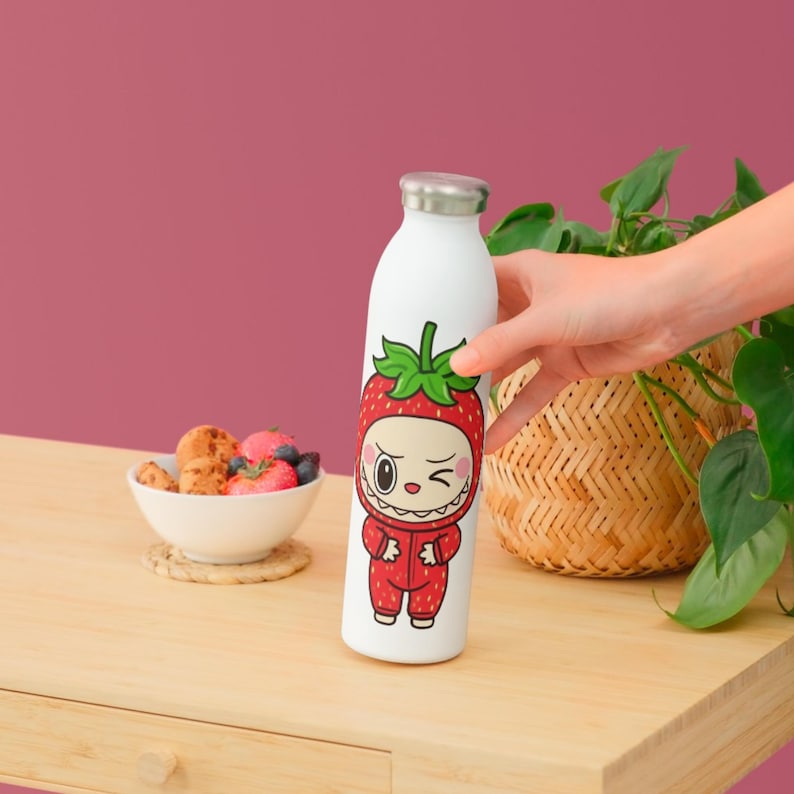 Puede incluir: Botella de agua blanca de acero inoxidable con tapa plateada. La botella presenta un personaje de fresa de dibujos animados con un mono rojo. El personaje tiene una expresi&oacute;n gui&ntilde;ando un ojo. La botella est&aacute; sujeta por una mano.