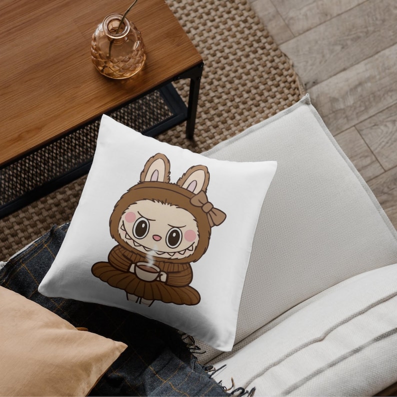 Puede incluir: Coj&iacute;n cuadrado blanco con un personaje de dibujos animados con un disfraz de conejo marr&oacute;n sosteniendo una taza de caf&eacute;. El personaje tiene ojos grandes y una expresi&oacute;n severa. El coj&iacute;n est&aacute; sobre un sof&aacute; con otros cojines.