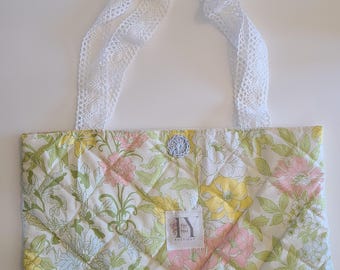 May include: A quilted tote bag with a floral pattern in yellow, pink, and green. The bag features white lace handles and a small, light blue crocheted embellishment. A label with the text "For You Boutique" is visible.