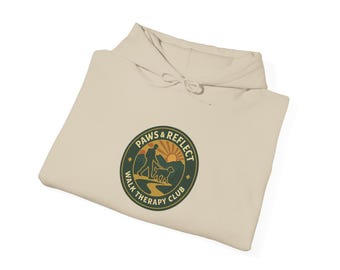 Puede incluir: Sudadera con capucha color crema con un emblema circular que dice "Paws & Reflect Walk Therapy Club". El emblema muestra a una persona paseando a un perro con una puesta de sol. La sudadera tiene una capucha con cordón.