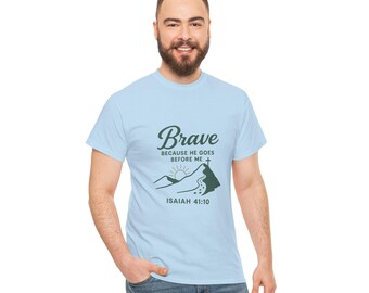 Può includere: T-shirt azzurra con la parola "Brave" in corsivo, insieme al testo "BECAUSE HE GOES BEFORE ME" e "ISAIAH 41:10". Il design include una scena di montagna con un sole e una croce. La maglietta è realizzata in materiale morbido.