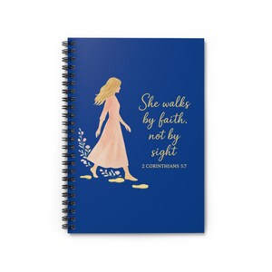 Puede incluir: Cuaderno de espiral azul marino con una ilustración de una mujer con un vestido melocotón. El texto "She walks by faith, not by sight" está escrito en oro, con el versículo bíblico 2 Corintios 5:7 debajo.