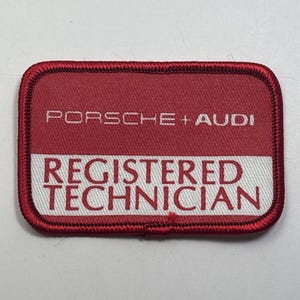 May include: A rectangular patch with rounded corners. The top section is red with the words "PORSCHE + AUDI" in white. The bottom section is white with the words "REGISTERED TECHNICIAN" in red. The patch has a red border.