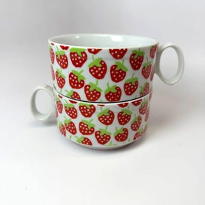 May include: Two white ceramic mugs stacked, each adorned with a repeating pattern of red strawberries with green leaves. The mugs have a rounded shape and a white handle. The background is plain white.