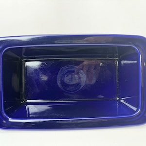 May include: A cobalt blue rectangular ceramic dish with rounded corners. The dish has a glossy finish and a recessed interior. The center of the dish features a circular design. The dish is empty and sits on a white surface.