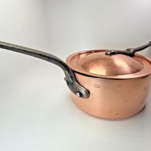 May include: A copper saucepan with a lid and a long, riveted handle. The handle and lid handle are dark gray. The copper pot has a shiny, reflective surface. The saucepan is a kitchenware item.