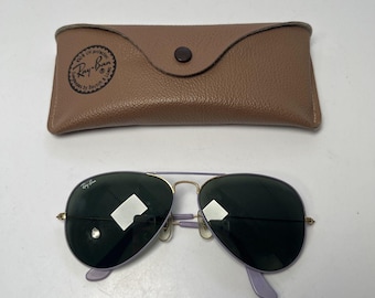 May include: Aviator-style sunglasses with a light purple frame and dark lenses, paired with a tan leather case. The sunglasses feature a gold bridge and temples, and the case has a button closure and a Ray-Ban logo.