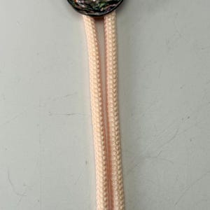 May include: A bolo tie with a round, iridescent abalone shell pendant. The pendant is attached to a light pink cord with silver-colored metal tips. The tie is a classic accessory.