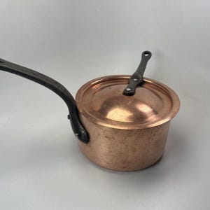 May include: A copper saucepan with a lid and a long, dark metal handle. The pot has a rounded shape and a matching lid with a small handle. The handle is riveted to the side of the pot. Kitchenware.