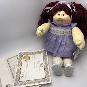 May include: A Cabbage Patch Kids doll with auburn yarn hair styled in pigtails, wearing a lavender dress with floral embroidery. The doll has a soft, plush body and is accompanied by birth certificates.