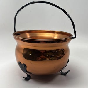 May include: A copper-colored cauldron with a black handle and three black feet. The cauldron has a rounded body and a wide opening. The handle is arched and attached to the sides of the cauldron. The feet are curved and provide stability.