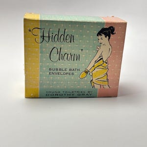 Könnte beinhalten: Ein rechteckiger Pappkarton für Hidden Charm Schaumbad-Umschläge von Dorothy Gray. Die Schachtel hat ein Farbschema in Hellblau, Gelb und Rosa mit einer grafischen Illustration einer Person, die in ein gelbes Handtuch gewickelt ist.