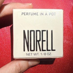 May include: A square, off-white cardboard box containing Norell perfume in a pot. The text on the front reads PERFUME IN A POT, NORELL, and NET WT. 1/8 OZ. in black lettering. The packaging is simple and features a clean, minimalist design.