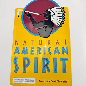 May include: Yellow metal sign with the text "NATURAL AMERICAN SPIRIT" in blue and red lettering. The sign features an illustration of a Native American figure smoking a pipe within a red and blue circle. The text "America's Best Cigarette" is also present.