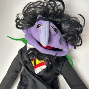 May include: A plush puppet of a cartoon character with a purple face, a long blue nose, and black curly hair. The puppet wears a black suit with a white, yellow, and red emblem. The puppet has large, round eyes and a red mouth.
